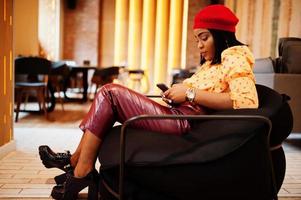 Elegant afro american woman in red french beret, big gold neck chain polka dot blouse and leather pants sit on pouf with mobile phone. photo