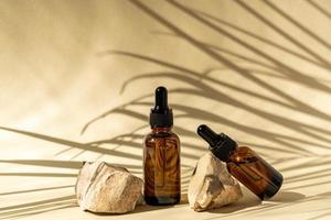 botellas cosméticas de vidrio oscuro con cuentagotas sobre un fondo beige con piedras y hojas tropicales. concepto de cosmética natural, aceites esenciales naturales y productos para el cuidado de la piel foto