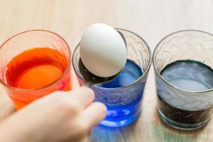 vasos de vidrio transparente con agua coloreada. el proceso de colorear los huevos. las manos de un niño se ponen en un huevo blanco con una cuchara. foto