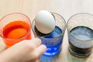Transparent glass cups with colored water. The process of coloring eggs. A child hands put in a white egg with a spoon. photo