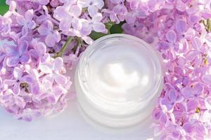 Top view of jar with face cream and lilac flowers on a light wooden background, place for text. Natural organic cosmetics concept. photo