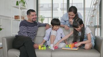 Happy Asian lovely Thai family care, dad, mum, and little children have fun playing with colorful toy blocks together on sofa in white living room, leisure weekend, and domestic wellbeing lifestyle. video
