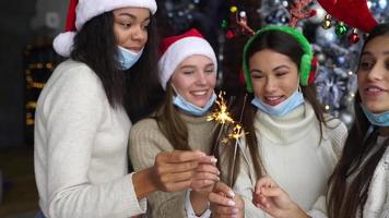 Young women at a holiday party wearing festive hats dance and laugh with sparklers in front of a Christmas tree video