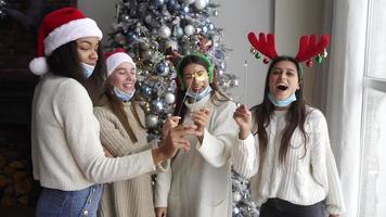 Young women at a holiday party wearing festive hats dance and laugh with sparklers in front of a Christmas tree video