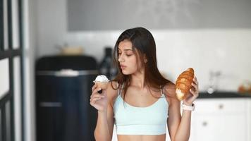 Young woman in active wear holds cupcake and croissant and look back and forth at both video