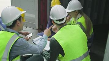 grupo de trabajadores multirraciales con uniformes de seguridad y cascos se sientan y discuten en pilas de contenedores logísticos con un gerente blanco masculino, intercambiando productos de envío para la industria del transporte de carga. video