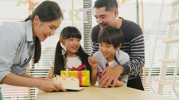 feliz familia asiática tailandesa, niños pequeños sorprendidos por pastel de cumpleaños, regalo, soplar una vela y celebrar la fiesta con padres y hermanos juntos en la mesa del comedor, evento especial de bienestar doméstico en el hogar. video