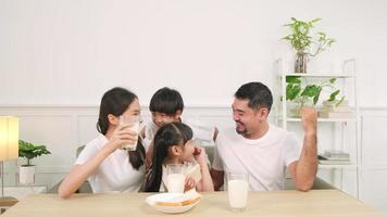 una familia tailandesa asiática saludable, niños pequeños y padres jóvenes beben leche blanca fresca en un vaso y pan de alegría juntos en una mesa de comedor en la mañana, bienestar nutrición hogar desayuno comida estilo de vida. video