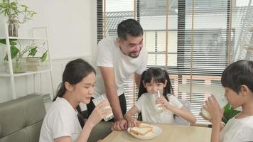 eine gesunde asiatische thailändische familie, kleine kinder und junge eltern trinken morgens frische weiße milch im glas und brotfreude zusammen an einem esstisch, wellness ernährung zuhause frühstücksmahlzeit lebensstil. video