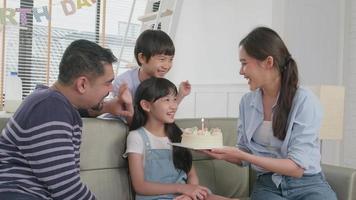 Happy Asian Thai family, young daughter is surprised with birthday cake, gift, blows out candle, and celebrates party with parents together in living room, wellbeing domestic home event lifestyle. video