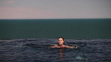 Beautiful woman swims in a pool of dark blue water and raises up at edge to look out video