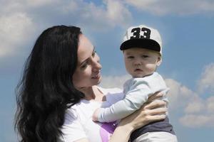 Happy young girl, mom plays with her baby in her arms against a background of blue sky with clouds. Copy space. photo