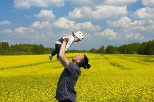 apuesto hombre de cabello oscuro, papá sostiene a su pequeño hijo en sus brazos en un campo de colza. copie el espacio foto