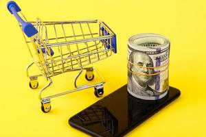 Mobile phone with a roll of one hundred dollar bills and a shopping cart on a yellow background. Online shopping concept. Copy space.