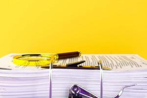 Close up of a pile of unfinished documents awaiting verification, a magnifying glass, on a yellow background. Business and education concept. Copy space photo