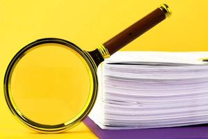 Close up of a pile of unfinished documents awaiting verification, a magnifying glass, on a yellow background. Business and education concept. Copy space photo