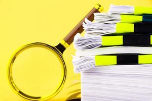 A large stack of unfinished documents awaiting verification, a magnifying glass, against a yellow background. Business and education concept. Copy space photo