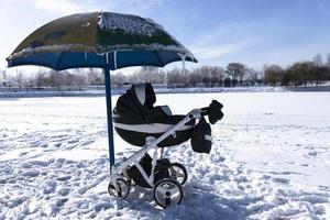 Baby carriage by the lake in winter. Beach under the snow and an umbrella from the sun. The concept of winter holidays with children. photo