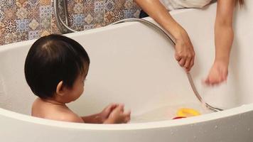 menino se divertindo tomando banho na banheira há uma mãe sentada ao lado