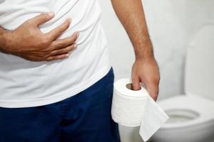 el hombre sufre de diarrea, sostenga el rollo de papel tisú frente a la taza del inodoro. estreñimiento en el baño. tratamiento dolor de estómago e higiene, cuidado de la salud foto