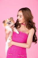 Girl wear chocky Pink dress hold cute dog and look at camera over pink background photo
