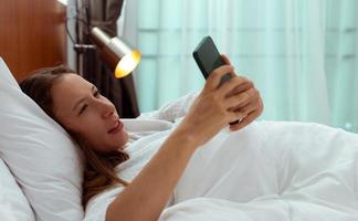 la mujer caucásica duerme en la almohada de la cama y la alarma de despertador del teléfono. las mujeres se sienten somnolientas y conmocionadas cuando toman un teléfono inteligente y ven la hora de trabajar al amanecer. adulto joven se siente inconsciente, copia espacio foto