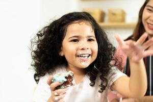 un niño en edad preescolar hace un pastel para cocinar en la cocina, una niña prepara una bonita decoración con diversión educada en cupcakes para la hermana de cumpleaños. desorden de crema en la cara y la boca del cabello rizado, siéntase feliz, copie el espacio foto