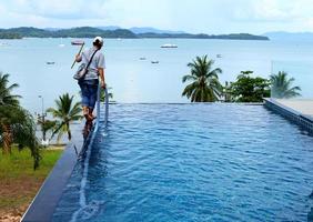 Woman Worker clean private swimming pool with vacuum blue tube cleaner every week in Summer, Salt type Pool along with Ocean as horizontal style with coconut trees photo