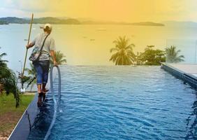 Woman Worker clean private swimming pool with vacuum blue tube cleaner every week in Summer, Salt type Pool along with Ocean as horizontal style with coconut trees
