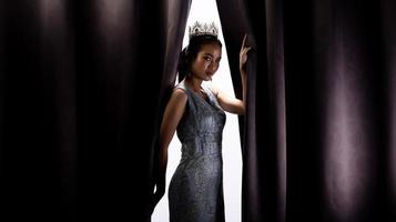 Miss Pageant Beauty Contest wears sequin Evening Gown long dress with sparkle Diamond Crown, Asian Woman opens purple Curtain after win Final round as new way life, opportunity, change, isolated photo