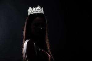 Portrait of Miss Pageant Beauty Contest in sequin Evening Ball Gown long dress with sparkle light Diamond Crown, silhouette low key exposure with curtain, studio lighting dark background dramatic photo