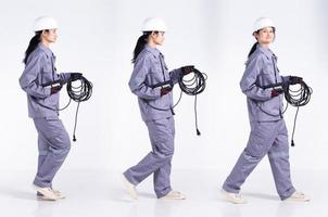 longitud completa 20s joven ingeniero de raza mixta mujer electricista, caminando hacia adelante izquierda derecha, usa uniforme gris. Contratista hembra mantenga cable de enchufe eléctrico sonrisa feliz sobre fondo blanco aislado foto