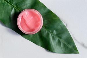 Pink body scrub on tropical leaf on marble background. Wellness and spa concept. Body care and treatment photo