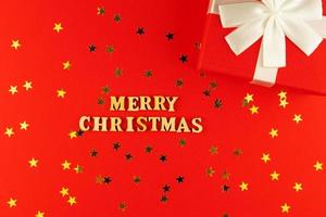 letras de madera sobre fondo rojo. letras de feliz navidad en papel rojo con confeti de estrellas dispersas y caja de regalo. foto