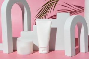 botellas de productos cosméticos en blanco sobre fondo rosa con rama de palma, como publicidad. tubo de plástico y tarro de crema junto a podios y arcos blancos. escaparate de moda foto