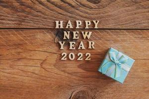 feliz año nuevo 2022. cita hecha de letras de madera y números 2022 sobre fondo de madera con caja de regalo pequeña azul. concepto creativo para la tarjeta de felicitación de año nuevo foto