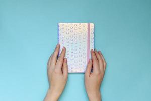 Female hands holding notebook with trendy holographic cover on blue background. Planning concept. Flat lay style. photo