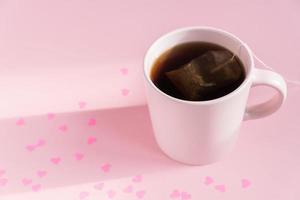 Pink cup with hot black tea on pink background with scattered paper hearts. Love and care concept. Copy space for text photo