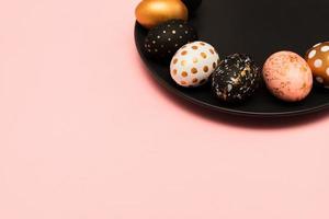 Pink background with white, black and golden decorated eggs on black plate. Trendy easter flat lay with copy space. photo