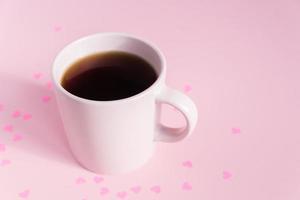 Pink cup with hot black tea on pink background with scattered paper hearts. Love and care concept. Copy space for text photo