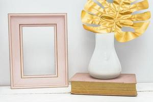 gold, pink and white interior mockup design. Empty photoframe and monstera leaf in white vase on vintage book on white wall background. Trendy design. photo