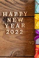 feliz año nuevo 2022. cita hecha de letras de madera y números 2022 sobre fondo de madera con pequeñas cajas de regalo multicolores. concepto creativo para la tarjeta de felicitación de año nuevo foto