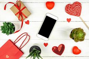 Creative Valentine Day romantic composition with red hearts, smartphone, lollipop, gift box and paper bag on pink background. Mockup with copy space for blogs and social media photo