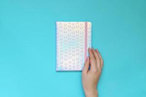 Female hand holding notebook with trendy holographic cover on blue background. Planning concept. Flat lay style. photo