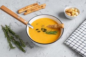 Pumpkin and carrot soup with cream, pumpkin seeds, croutons and grossini bread sticks on dar k wooden background. Rustic style, selective focus photo