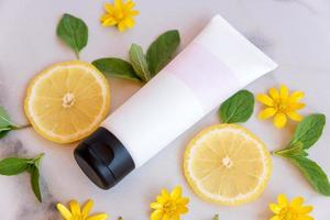 Top view facial AHA skincare product. white tube with blank label decorated with fresh lemon slices and green leaves on marbe background. Refresh skin Vitamin c concept photo