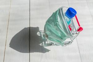 Shopping cart in medical mask with bottle of sanitizer on wgite wooden background. Coronavirus prevention concept. Top view. photo