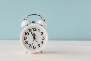White alarm clock standing on wooden shelf on blue background. copy space fro your text or design. Fime minutes before twelve on a display photo