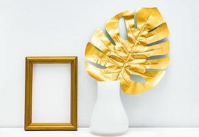 diseño de maqueta interior en oro y blanco. marco de fotos vacío y hoja de monstera en un jarrón blanco sobre fondo de pared blanca. diseño de moda