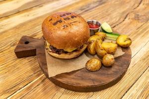 hamburguesa con carne servida con pepinos, cetchup y patata frita en tabla de cortar redonda. cena poco saludable foto